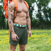 Shirtless man standing in forest wearing hiking backpack and olive green lounge shorts.
