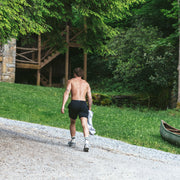 Shirtless man walking in forest with canoe and cabin in background wearing black lounge shorts.