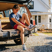 Man sitting in truck of jeep wearing blue shirt, red shorts, and putting on blue performance ankle socks.