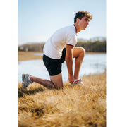 Man in field adjusting shoe wearing shite shirt and black shorts.