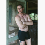 Shirtless man with arms crossed standing in front of open cabin door wearing black boxer briefs.