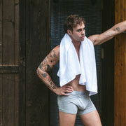 Shirtless man standing outside of an outdoor shower with towel over shoulder wearing grey boxer briefs.