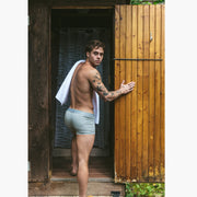 Back shot of shirtless man walking into outdoor shower with towel on shoulder wearing grey boxer briefs.