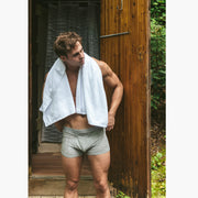 Shirtless man standing outside of outdoor shower with towel on shoulder wearing grey boxer briefs.