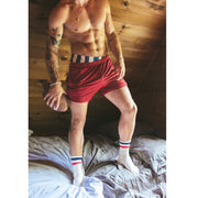Man standing on bed shirtless holding a football and wearing burgundy mesh lounge shorts.