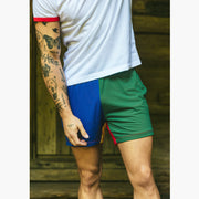 Close up of man standing in front of cabin wearing white t-shirt and blue/gree/yellow/red colored mesh lounge shorts.