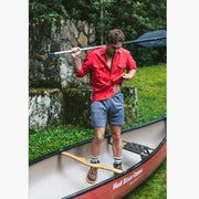 Man standing inside of canoe with paddle over shoulder weariing red shirt and grey lounge shorts.