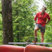 Man in forest standing on stack of canoes wearing a red shirt and grey lounge shorts.
