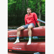 Man in forest sitting on top of pile of canoes wearing red shirt and grey lounge shorts.