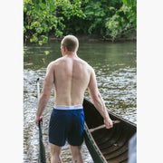 Back shot of a shirtless man pulling a canoe in water holding a paddle and wearing navy blue lounge shorts.