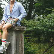 Man sitting in forest on cement pillar wearing Madras print lounge short, blue opened dress shirt, and white/red/blue crew socks.