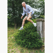 Man in forest jumping off cement pillar wearing blue dress shirt, madras printed lounge short, and red/white/blue crew socks.