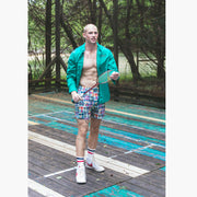 Full body shot of man wearing green dress shirt and Madras print lounge shorts standing on wooden court holding a racket.