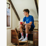 Man sitting on desk wearing blue shirt and white crew socks with letters "nila" in red.