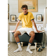 Man sitting on edge of bath wearing yellow shirt, white shorts and white/yellow/blue crew socks with letters NILA on them