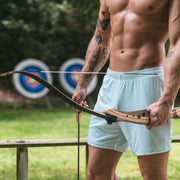 Shirtless man standing in archery range holding a bow and wearing white/green mesh lounge shorts.