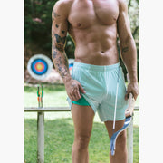 Shirtless man standing in archery range holding a bow and wearing white/green mesh lounge shorts. The man is lifting the shorts to show the green interior.