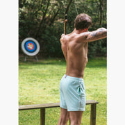 Back shot of shirtless man standing in archery range holding a bow aiming it at target and wearing white/green mesh lounge shorts.