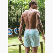 Back shot of shirtless man standing at archery range holding a box wearing white mesh shorts.