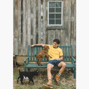 Man sitting on bench with small goat. Man is wearing yellow shirt, boots and blue slim fit boxers with white dots.