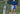 Man standing in bushes by beach wearing blue sweater and checkered/solid navy blue lounge shorts.
