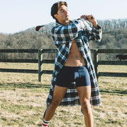 Shirtless man wearing open robe swinging a golf club outside with trees in background. He is also wearing blue boxer briefs.