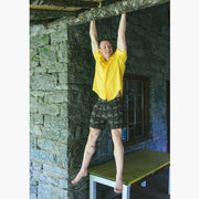 Man hanging in the air holding on to a wooden beam wearing a yellow polo and black "schooner" mesh lounge shorts.