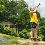 Man at camp playing a trumpet wearing yellow shirt and blue mesh lounge shorts with merit badges on them. 