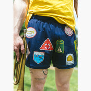 Close up shot of man holding trumpet and wearing yellow shirt with blue mesh lounge shorts that have merit badges on them.