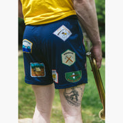 Close up back shot of man holding trumpet and wearing yellow shirt with blue mesh lounge shorts that have merit badges on them.