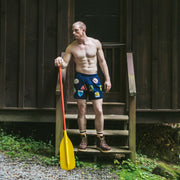 Shirtless man holding yellow paddle wearing blue mesh lounge shorts with merit badges printed on them.