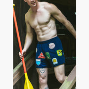 Shirtless man holding a yellow paddle wearing blue mesh lounge shorts with merit badges printed on them.