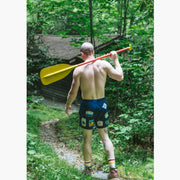 Back shot of shirtless man holding yellow paddle wearing blue mesh lounge shorts with merit badges printed on them.